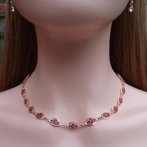 Könnte beinhalten: Ein goldfarbenes Halsketten- und Ohrring-Set. Die Halskette hat oval geformte rubinrote Steine und kleine klare Steine. Die Ohrringe haben ein ähnliches Design mit rubinroten und klaren Steinen.