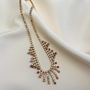 May include: A gold-toned necklace with iridescent rhinestones. The necklace features a delicate chain and a striking pendant design with a sunburst-like arrangement of colorful stones. The stones are in shades of red, pink, and blue.