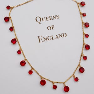 以下が含まれることがあります： 複数の赤い丸い宝石が付いたゴールドカラーのネックレス。 ネックレスは繊細なチェーンと留め金が付いています。 背景には「QUEENS OF ENGLAND」の文字が印刷されています。