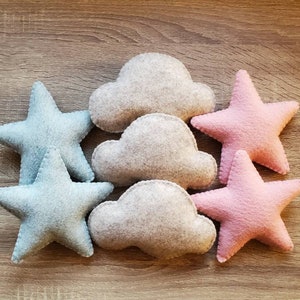 Puede incluir: Seis decoraciones de fieltro en forma de estrellas y nubes. Dos de las estrellas son azul claro, dos son rosas y dos de las nubes son grises.