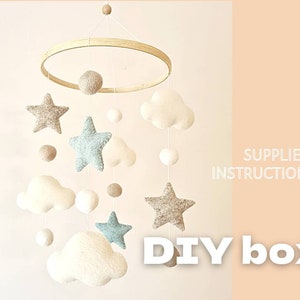 Può includere: Mobile per neonati fatto a mano con stelle, nuvole e sfere in feltro dai colori neutri. Sospeso a un cerchio di legno. Il testo "DIY box" e "SUPPLIES + INSTRUCTIONS" sono visibili.
