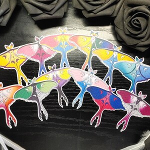 Puede incluir: Un conjunto de nueve pegatinas de polillas coloridas con un diseño de luna creciente. Las polillas están dispuestas en semicírculo y tienen una variedad de colores, incluyendo amarillo, rosa, azul, verde y morado.