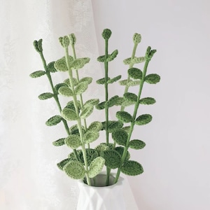 May include: A white ceramic vase holds a collection of handmade crochet stems with green leaves. The stems are a light green color, and the leaves are a darker shade of green. The vase has a geometric design.