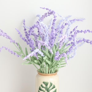 Puede incluir: Un ramo de flores de lavanda de ganchillo en tonos de morado y azul claro, dispuestas en un jarrón de cerámica crema con un diseño de hoja de monstera verde.