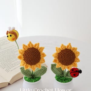 May include: Crocheted sunflower and bee decorations in white pots. The sunflowers have yellow petals and brown centers. A red and black ladybug is attached to one. The text "Little Crochet Flower" is visible.
