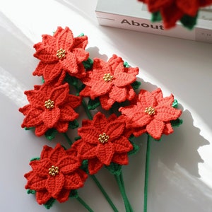 Puede incluir: Un ramo de seis flores de poinsettia de crochet rojo con centros de cuentas doradas y tallos verdes.