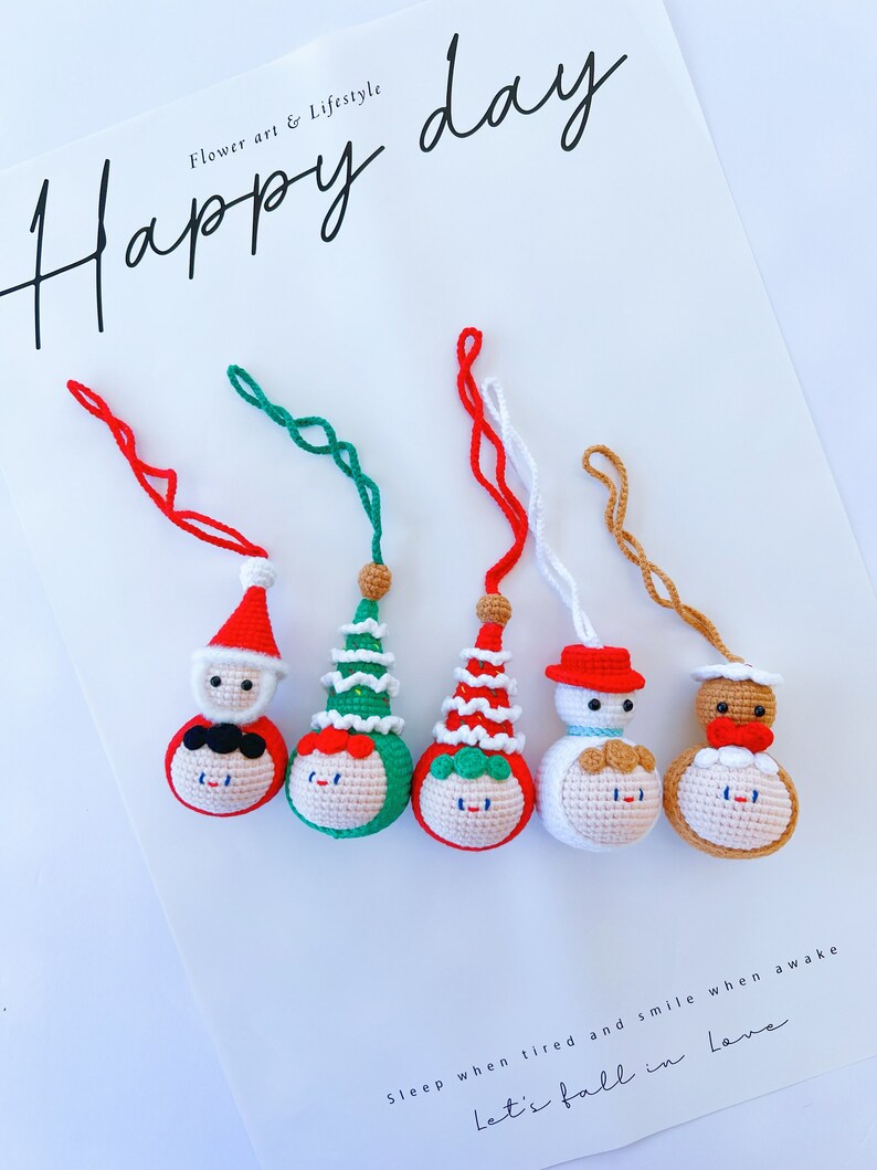 May include: Five crocheted ornaments with different festive themes. The ornaments are red, green, white, and brown. Each ornament has a smiling face and a different hat. The ornaments are hanging from red, green, white, and brown strings. The text "Happy day" is written in black at the top of the image. The text "Sleep when tired and smile when awake Let's fall in love" is written in black at the bottom of the image.