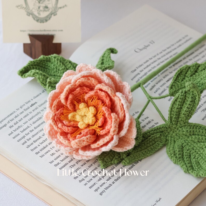 K&ouml;nnte beinhalten: Eine handgefertigte H&auml;kelblume mit pfirsich- und orangefarbenen Bl&uuml;tenbl&auml;ttern und einem gelben Zentrum. Die Blume liegt auf einem offenen Buch mit gr&uuml;nen H&auml;kelbl&auml;ttern und -stiel. Ein einzigartiger, handgefertigter Artikel.