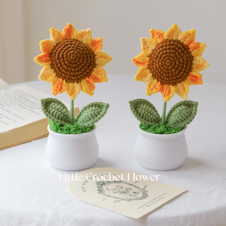 May include: Two handmade crochet sunflowers in white ceramic pots. The sunflowers have yellow and orange petals with brown centers. Green leaves and moss-like material surround the stems. The text "Little Crochet Flower" is visible.