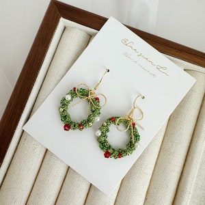 May include: A pair of green and red Christmas wreath earrings with white pearl beads and gold accents. The earrings are handmade and feature a small bow on each side. The earrings are displayed on a white card with the text "Hua Jian Li Hand crochet".