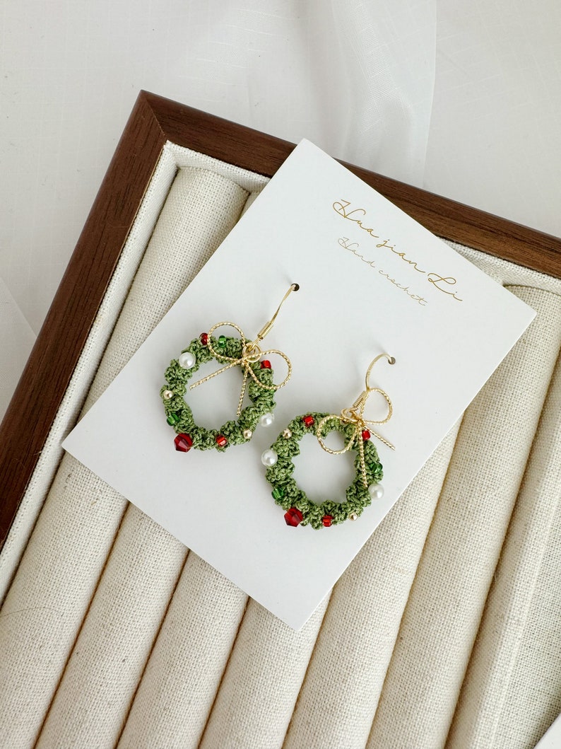 May include: A pair of green and red Christmas wreath earrings with white pearl beads and gold accents. The earrings are handmade and feature a small bow on each side. The earrings are displayed on a white card with the text "Hua Jian Li Hand crochet".