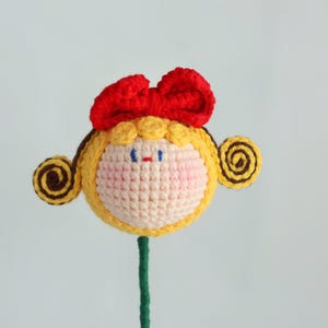 Puede incluir: Una cabeza de mu&ntilde;eca de ganchillo hecha a mano con un lazo rojo, pelo amarillo y un tallo verde. La cara de la mu&ntilde;eca es de color crema con mejillas rosadas, ojos azules y boca roja. El pelo est&aacute; peinado con rizos en espiral marrones a cada lado.