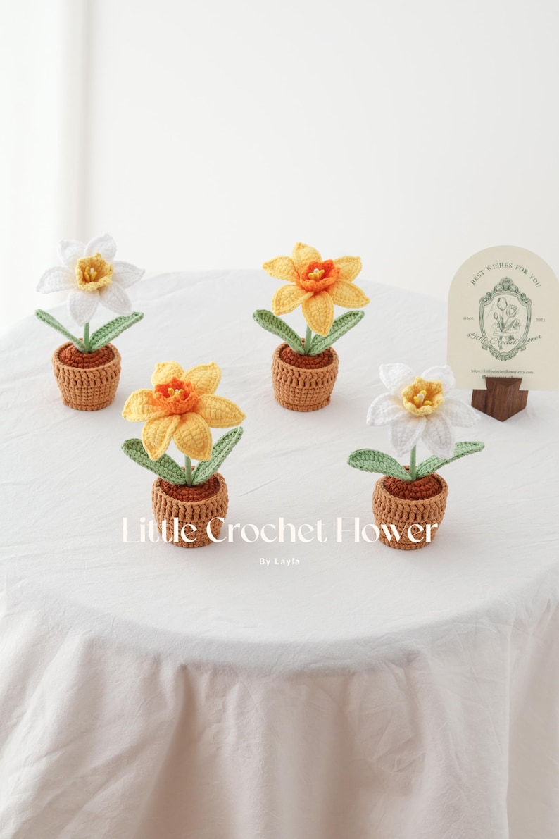 K&ouml;nnte beinhalten: Geh&auml;kelte Narzissen in kleinen braunen T&ouml;pfen. Die Blumen sind gelb und wei&szlig; mit gr&uuml;nen Bl&auml;ttern. Das Bild enth&auml;lt eine Karte mit der Aufschrift "Best Wishes For You". Der Text "Little Crochet Flower" ist ebenfalls im Bild enthalten.