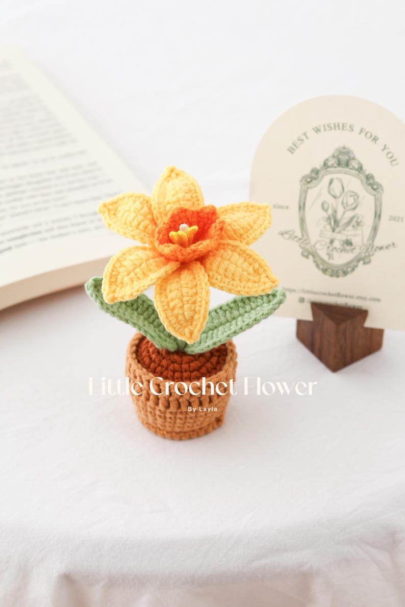 K&ouml;nnte beinhalten: Geh&auml;kelte Narzissenblume in einem kleinen braunen Topf. Die Blume hat gelbe Bl&uuml;tenbl&auml;tter mit einem orangefarbenen Zentrum und gr&uuml;nen Bl&auml;ttern. Der Text "Little Crochet Flower" ist sichtbar.