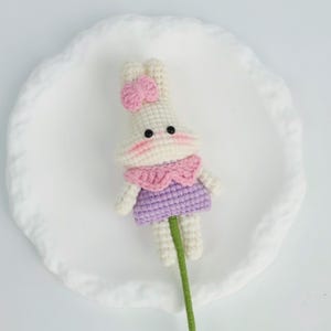 Puede incluir: Mu&ntilde;eca de conejo hecha a mano con un lazo rosa, un cuello rosa y un vestido morado. El conejo tiene ojos negros y mejillas rosas. Est&aacute; unido a un tallo verde, hecho a mano.