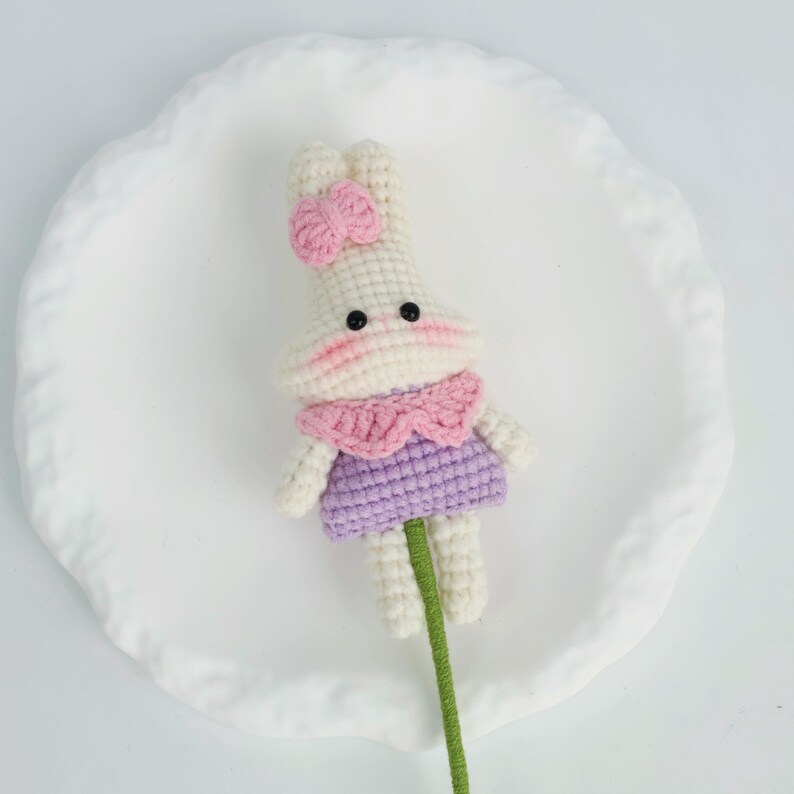 Puede incluir: Mu&ntilde;eca de conejo hecha a mano con un lazo rosa, un cuello rosa y un vestido morado. El conejo tiene ojos negros y mejillas rosas. Est&aacute; unido a un tallo verde, hecho a mano.
