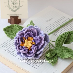 K&ouml;nnte beinhalten: Eine handgefertigte H&auml;kelblume mit einem lila und gelben Zentrum, gr&uuml;nen Bl&auml;ttern und einem gr&uuml;nen Stiel. Die Blume liegt auf einem offenen Buch. Der Text "little crochet flower" ist sichtbar.