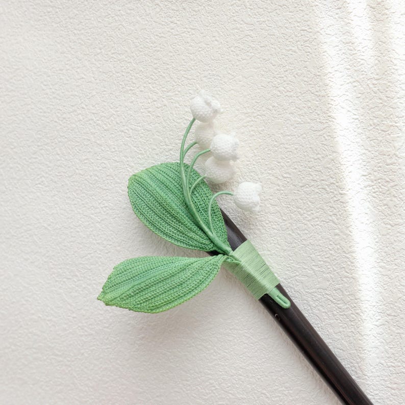 May include: A handcrafted hair stick featuring a lily of the valley design. The stick has a dark brown wooden base, with green crocheted leaves and stems, and white crocheted flower buds. The design is secured with green thread.