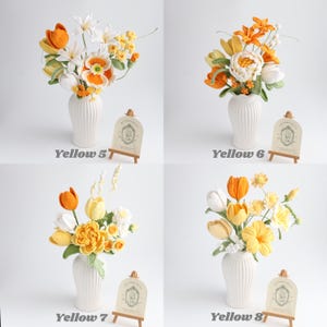 May include: Four arrangements of crocheted flowers in white fluted vases, labeled Yellow 5 through Yellow 8. Each bouquet features various shades of yellow, orange, and white yarn blossoms with green stems, accompanied by a small wooden easel sign.