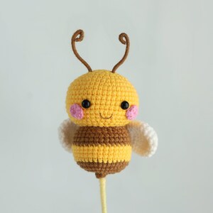 Puede incluir: Un juguete de abeja hecho a mano con ganchillo. La abeja tiene una cabeza amarilla con mejillas rosas, ojos negros y antenas marrones. El cuerpo es de rayas amarillas y marrones, con alas blancas. La abeja est&aacute; unida a un palo amarillo.