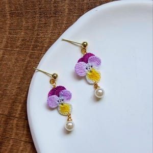 May include: A pair of crocheted flower earrings with a pearl accent. The flowers are purple, pink, and yellow.