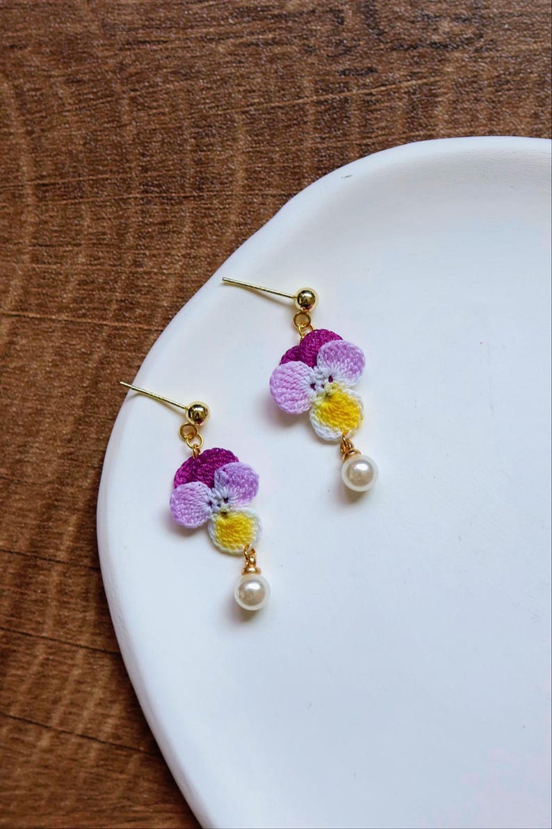 May include: A pair of crocheted flower earrings with a pearl accent. The flowers are purple, pink, and yellow.