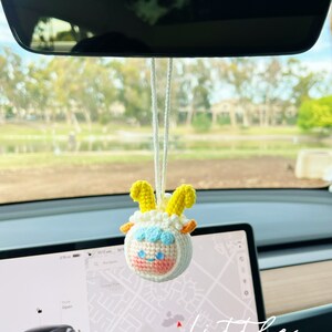 Puede incluir: Una oveja de crochet blanca con cuernos amarillos y una cara rosa colgando del espejo retrovisor de un coche. La oveja lleva un gorro de crochet azul y blanco. La palabra "little" est&aacute; escrita en blanco sobre un fondo negro en la esquina inferior derecha de la imagen.