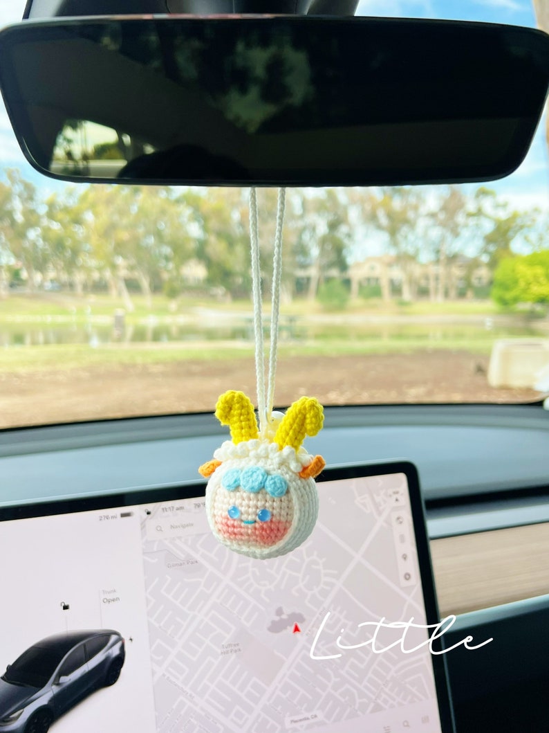 Puede incluir: Una oveja de crochet blanca con cuernos amarillos y una cara rosa colgando del espejo retrovisor de un coche. La oveja lleva un gorro de crochet azul y blanco. La palabra "little" est&aacute; escrita en blanco sobre un fondo negro en la esquina inferior derecha de la imagen.
