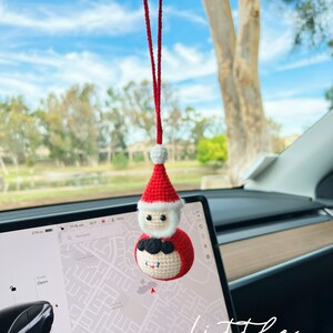 May include: A red and white crocheted Santa Claus ornament hanging from the rearview mirror of a car. The ornament is a round ball with a Santa Claus face and a white beard. The ornament is hanging from a red string.