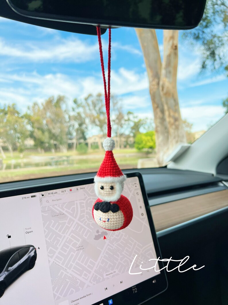 May include: A red and white crocheted Santa Claus ornament hanging from the rearview mirror of a car. The ornament is a round ball with a Santa Claus face and a white beard. The ornament is hanging from a red string.