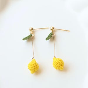 Puede incluir: Un par de pendientes de lim&oacute;n amarillo hechos a crochet con hojas verdes de crochet que cuelgan de postes dorados. Los pendientes est&aacute;n sobre un fondo blanco. Little Crochet Flower By Layla.