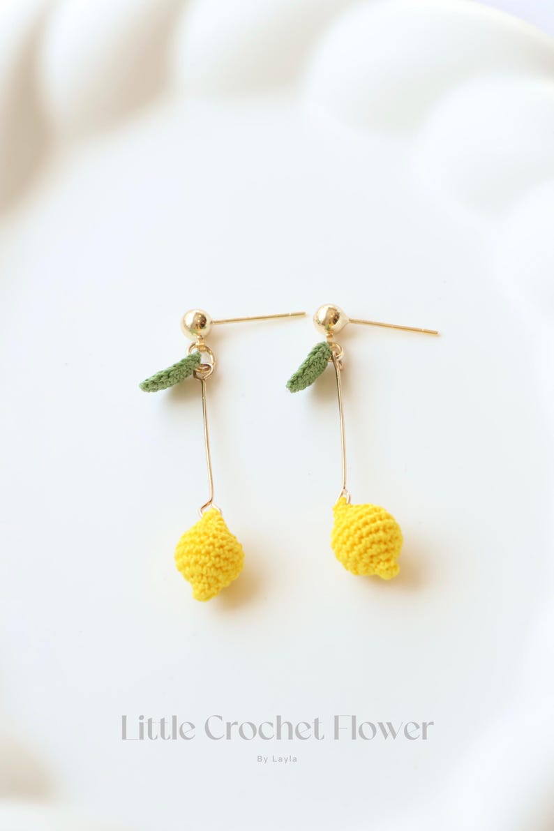 Puede incluir: Un par de pendientes de lim&oacute;n amarillo hechos a crochet con hojas verdes de crochet que cuelgan de postes dorados. Los pendientes est&aacute;n sobre un fondo blanco. Little Crochet Flower By Layla.