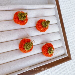May include: Four crocheted pumpkin earrings with gold hooks. The pumpkins are orange with green stems and are displayed in a white and brown jewelry box.