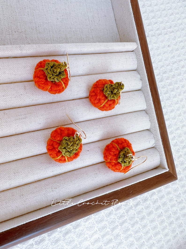 May include: Four crocheted pumpkin earrings with gold hooks. The pumpkins are orange with green stems and are displayed in a white and brown jewelry box.