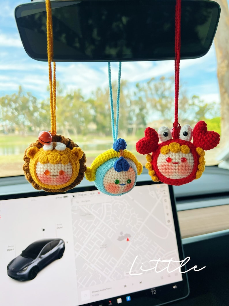 Puede incluir: Tres figuras de animales de crochet colgando del espejo retrovisor de un coche. Las figuras son un le&oacute;n amarillo, un oso azul y blanco y un cangrejo rojo. Las figuras son todas sonrientes y tienen ojos grandes.