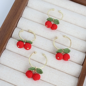 May include: Four gold-toned adjustable rings, each adorned with a pair of red crocheted cherries and green leaves. The rings are displayed on a white fabric surface within a wooden jewelry box.