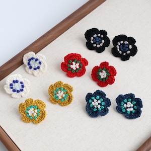 May include: A collection of handmade crochet flower earrings in various colors, including white, red, yellow, navy blue, and black. Each flower has a contrasting center, and the earrings are displayed on a neutral surface.