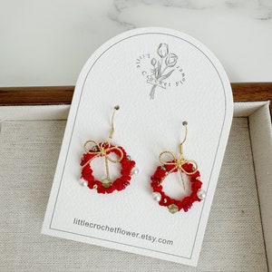May include: Red crocheted Christmas wreath earrings with gold wire and white pearl accents. The earrings are displayed on a white card with the text "littlecrochetflower.etsy.com".
