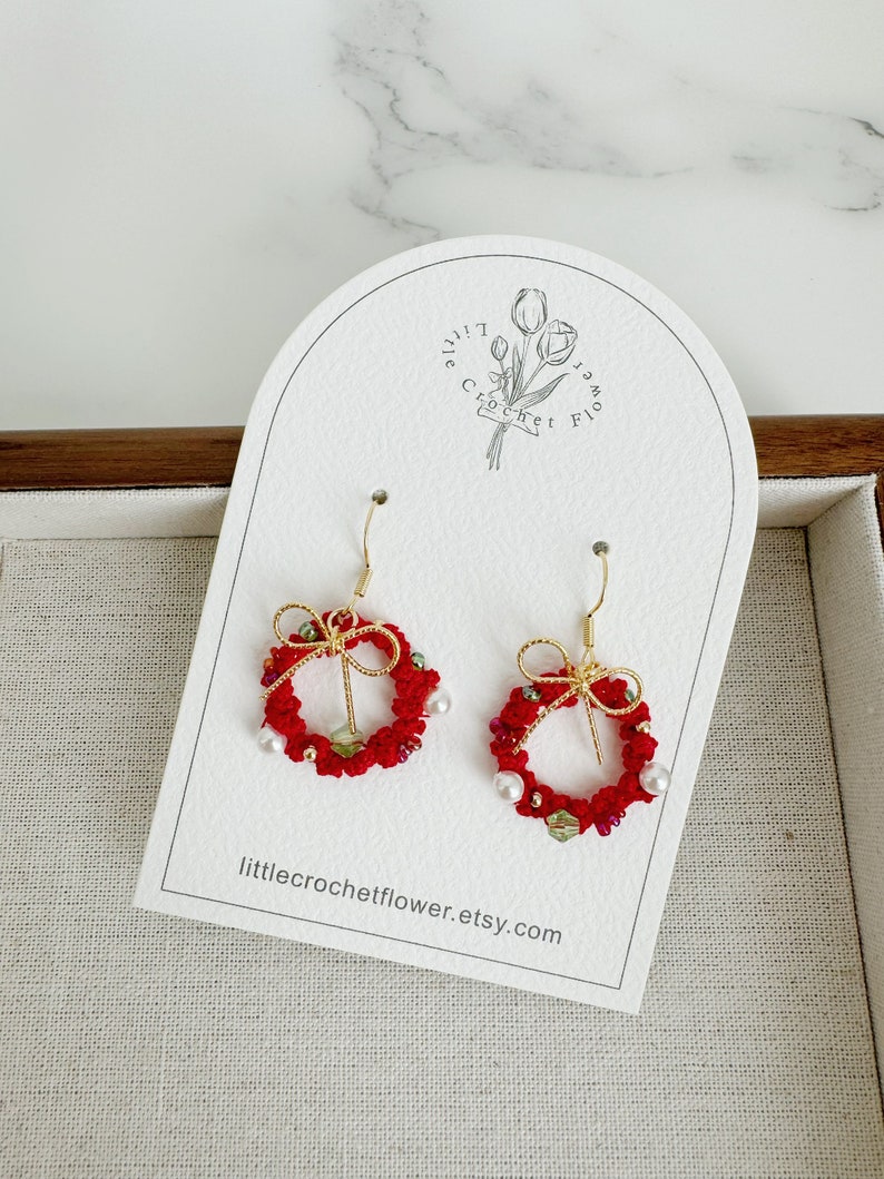 May include: Red crocheted Christmas wreath earrings with gold wire and white pearl accents. The earrings are displayed on a white card with the text "littlecrochetflower.etsy.com".