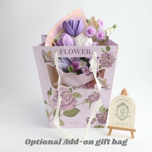 May include: A light purple gift bag featuring floral illustrations, butterfly graphics, and the word FLOWER, containing a bouquet of crocheted purple and white flowers. Beside it stands a small wooden easel holding a card with floral line art.