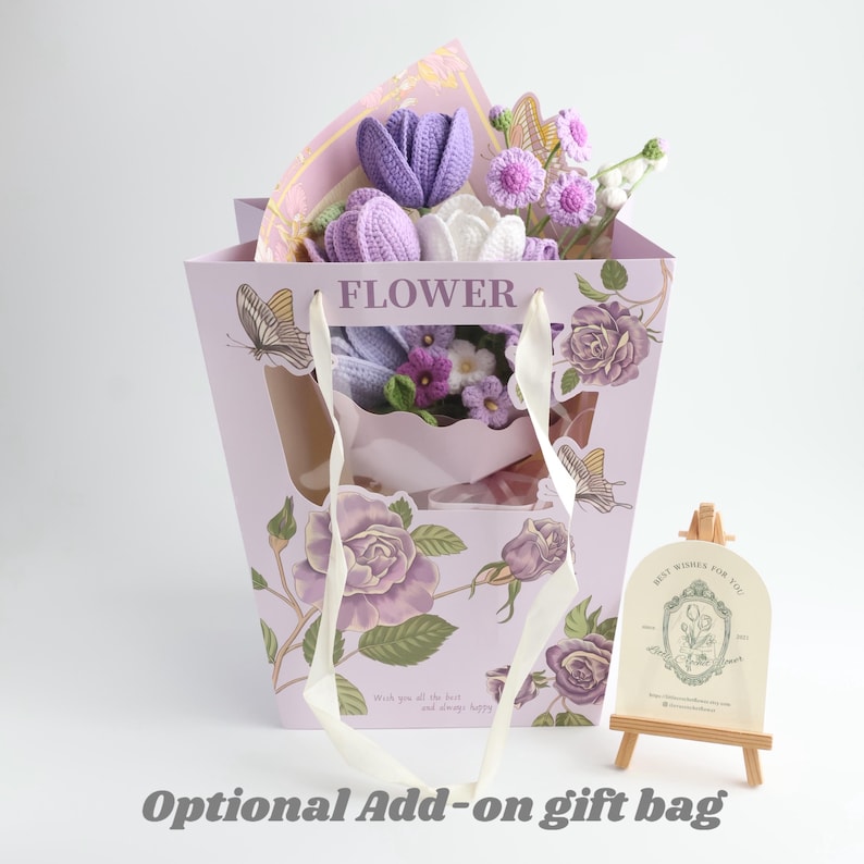 May include: A light purple gift bag featuring floral illustrations, butterfly graphics, and the word FLOWER, containing a bouquet of crocheted purple and white flowers. Beside it stands a small wooden easel holding a card with floral line art.