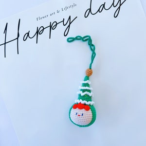 May include: A green crocheted Christmas tree ornament with a smiling face and a white crocheted hat. The ornament is hanging from a green string. The text "Happy day" is written in black cursive on a white background.