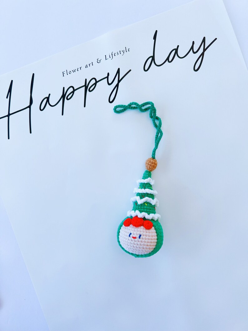 May include: A green crocheted Christmas tree ornament with a smiling face and a white crocheted hat. The ornament is hanging from a green string. The text "Happy day" is written in black cursive on a white background.