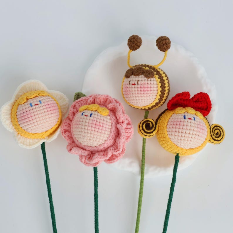 Puede incluir: Cuatro figuras de ganchillo hechas a mano de flores e insectos. Las figuras incluyen dos flores, una abeja y un personaje con un lazo rojo. Cada figura tiene una cara y est&aacute; unida a un tallo verde. Las flores son amarillas y rosas.