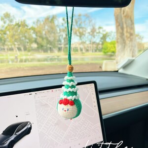May include: A green and white crocheted Christmas tree ornament with a red face and a white beard hanging from the rearview mirror of a car. The word "little" is written in white on the dashboard of the car.