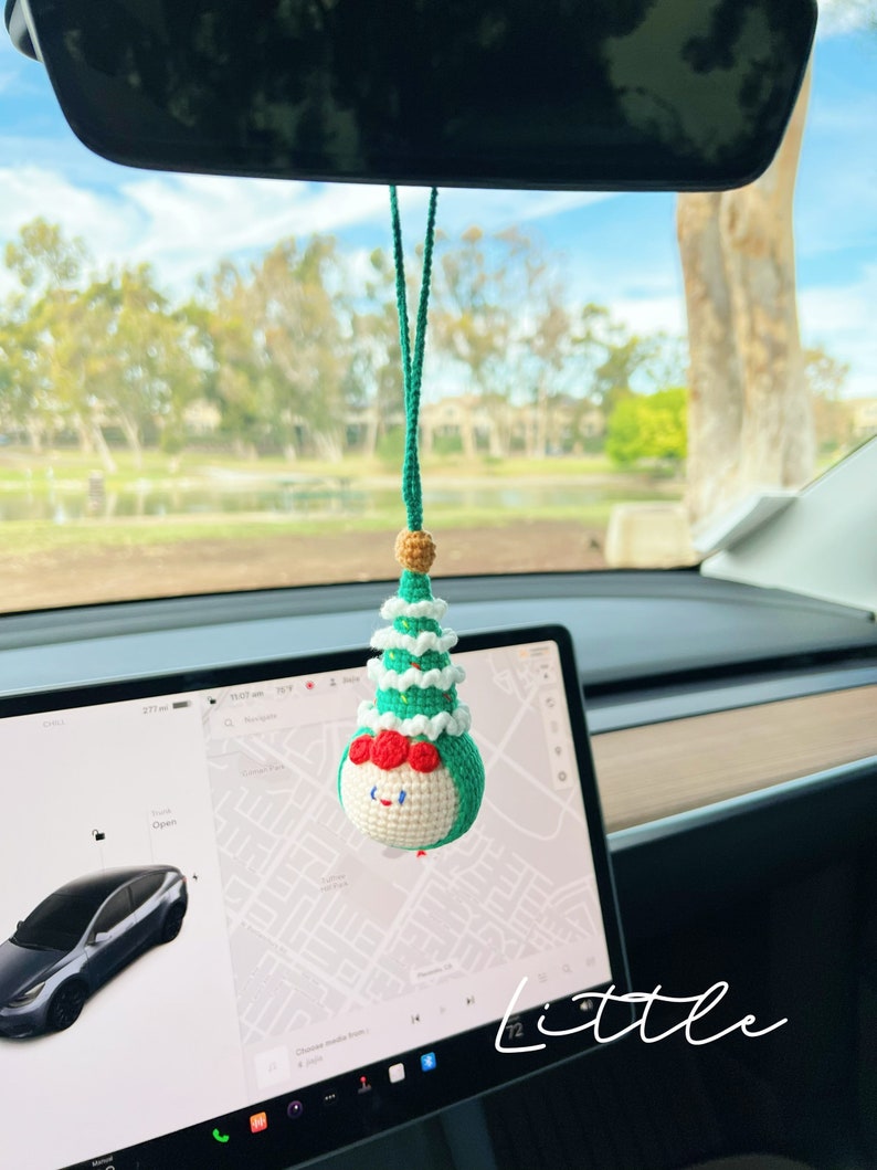 May include: A green and white crocheted Christmas tree ornament with a red face and a white beard hanging from the rearview mirror of a car. The word "little" is written in white on the dashboard of the car.