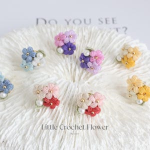 May include: Assortment of colorful crocheted flower rings with pearl accents. The rings are displayed on a white coral-like surface. The text "Little Crochet Flower" is written below the rings.