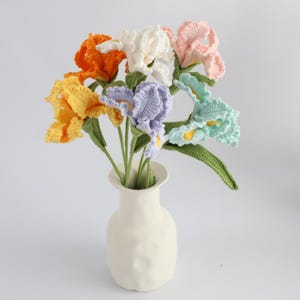 May include: A bouquet of six crocheted iris flowers in orange, yellow, white, light purple, light pink, and mint green, each with green stems and leaves, arranged in a matte white ceramic vase with a textured, organic, rounded shape.