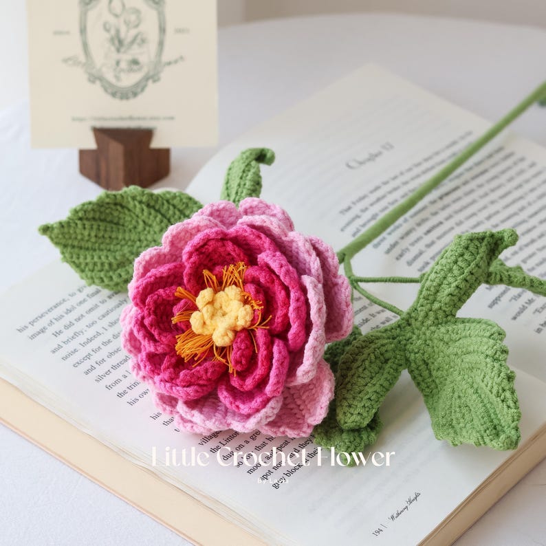 K&ouml;nnte beinhalten: Eine handgefertigte H&auml;kelblume mit rosa und magenta Bl&uuml;tenbl&auml;ttern, einem gelben Zentrum und gr&uuml;nen Bl&auml;ttern und Stiel. Die Blume liegt auf einem offenen Buch. Der Text "Little crochet Flower" ist sichtbar.