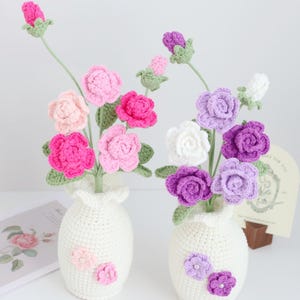 May include: Two white crocheted vases with crocheted flowers. The vase on the left has pink flowers and the vase on the right has purple and white flowers. The vases are decorated with small crocheted flowers.